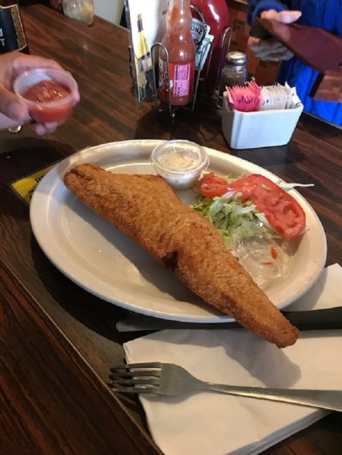 A large, golden-brown fried fish fillet on a plate with lettuce, tomato, and dipping sauces.