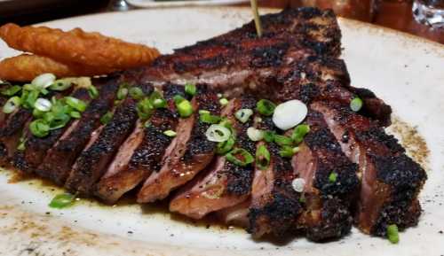 A plate of sliced, grilled steak topped with green onions, accompanied by two crispy potato wedges.