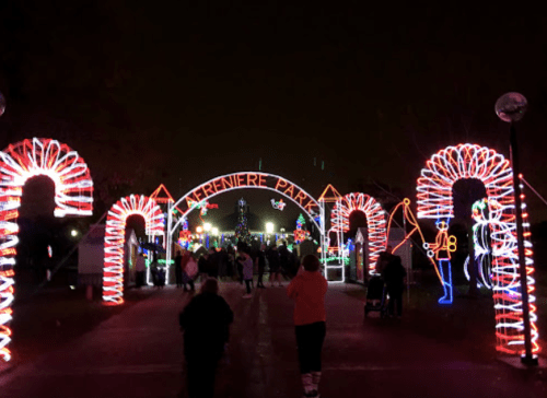 Colorful holiday lights illuminate a park entrance, with candy cane arches and festive decorations, as visitors stroll through.