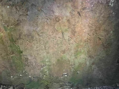 Ancient stone wall with carved symbols and markings, partially covered in moss and dirt.