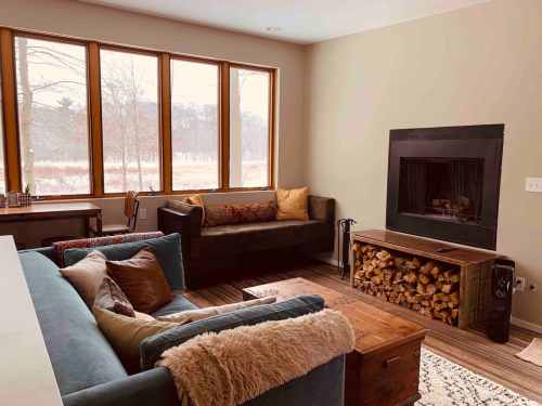 Cozy living room with large windows, a fireplace, and comfortable seating, featuring a wooden coffee table and decorative pillows.