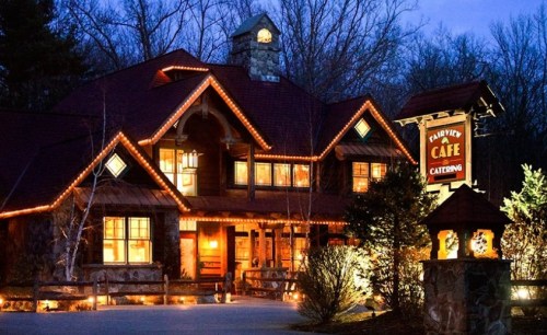 A cozy café with warm lights, surrounded by trees, set against a twilight sky. The building features rustic architecture.