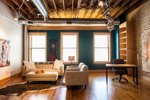 Modern loft-style living space with a sofa, desk, and large windows, featuring wooden beams and colorful wall art.