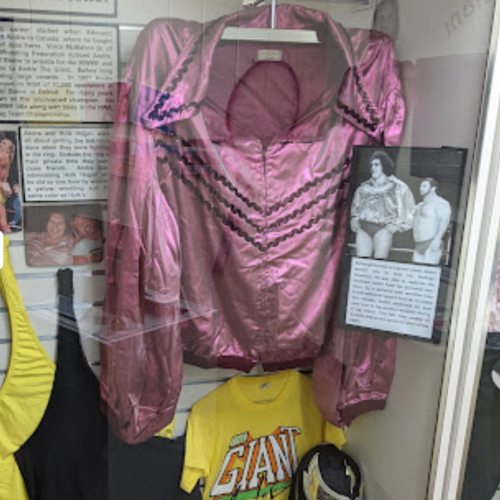 A pink satin shirt displayed in a glass case, alongside a yellow shirt and informational plaques about wrestling history.