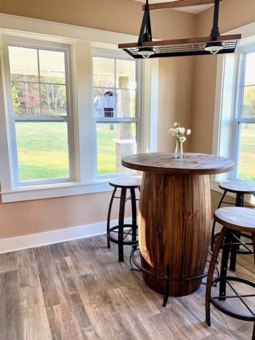 A wooden barrel table with three stools, set by large windows overlooking a green landscape.