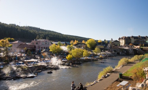 A scenic riverside view with steam rising, surrounded by trees and buildings under a clear blue sky.