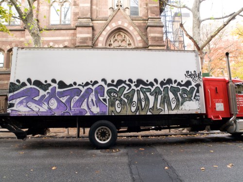 A white truck with purple graffiti on the side, parked on a city street near a historic building.