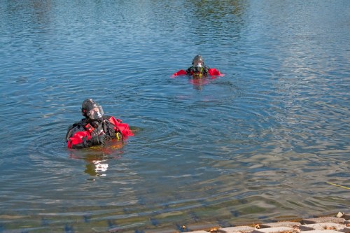 Two divers in red suits and helmets are swimming in a calm, clear body of water.