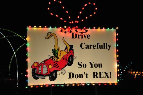 A colorful sign with a cartoon dinosaur in a car, urging drivers to "Drive Carefully So You Don't REX!"