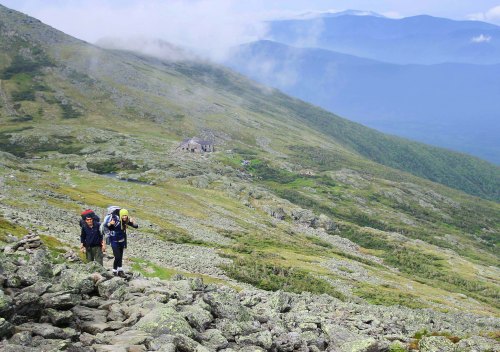 Two hikers ascend a rocky mountain trail, with a distant building and misty mountains in the background.