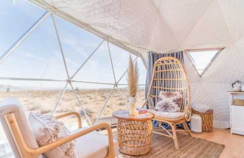 Cozy interior of a geodesic dome with a rattan chair, small table, and desert view through large windows.