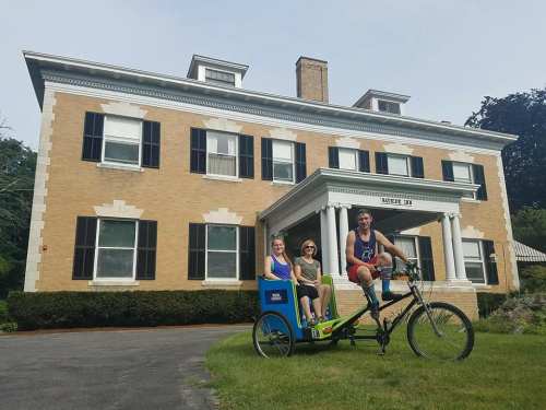 Three people pose on a bicycle rickshaw in front of a large, historic brick house with black shutters.