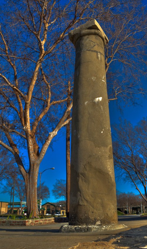 A tall, weathered stone column stands beside a bare tree against a blue sky in a park setting.