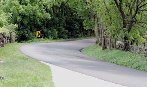 A winding road curves through a lush green landscape with trees lining the sides. A caution sign indicates a turn ahead.
