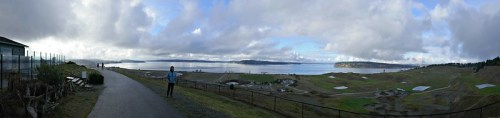 A panoramic view of a coastal landscape with a path, water, and distant hills under a cloudy sky.