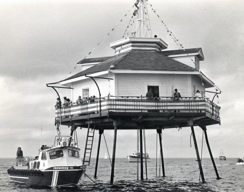 A historic seaside structure on stilts with people on the deck, alongside a small boat named "Frederick."
