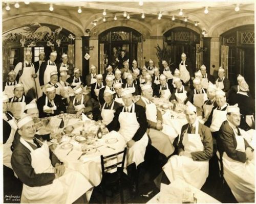 A large group of men in white aprons and hats gathered around a dining table in a vintage restaurant setting.