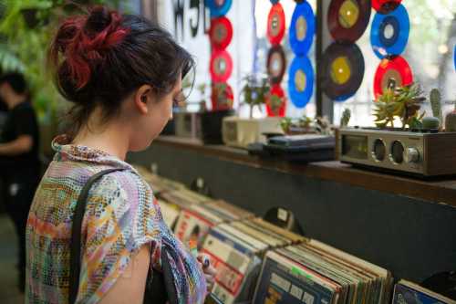 A person with colorful hair browses vinyl records in a cozy shop, surrounded by plants and vintage decor.