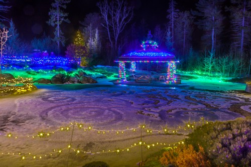 A winter scene featuring a gazebo adorned with colorful lights, surrounded by illuminated trees and a snowy landscape.