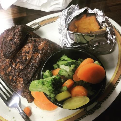A plate with grilled meat, a baked sweet potato wrapped in foil, and a side of mixed vegetables including broccoli and carrots.