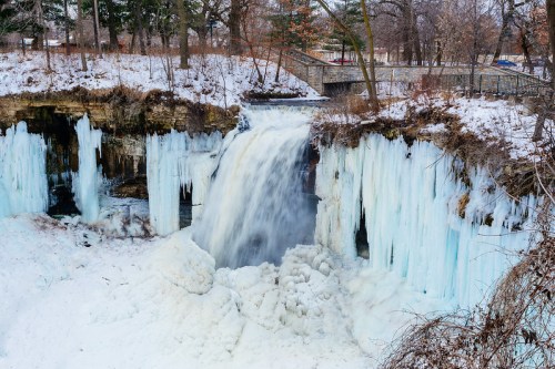 A frozen waterfall surrounded by icy formations and snow, with trees and a pathway in the background.