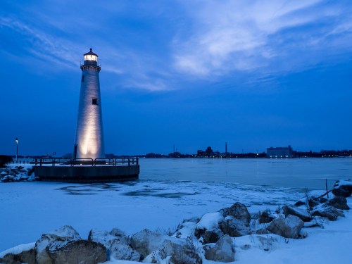 A lighthouse stands by a frozen lake at dusk, surrounded by snow and rocks, under a blue sky.