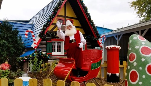 A cheerful Santa Claus waves from a red sleigh in front of a festive log cabin decorated for Christmas.