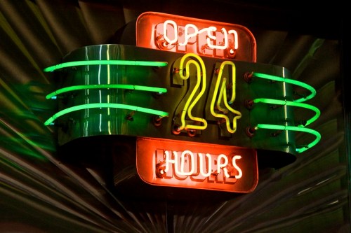 Neon sign reading "Open 24 Hours" with vibrant green and orange lights against a dark background.