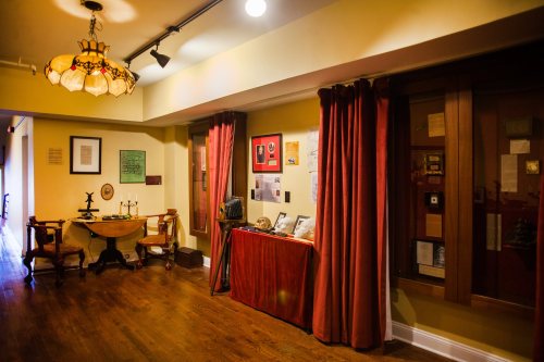 A warmly lit room featuring a wooden table, red curtains, and display cases with historical artifacts and documents.
