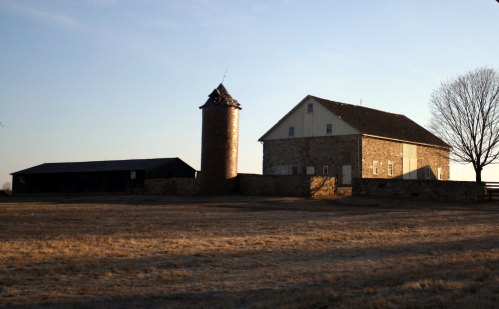 A rustic farm scene featuring a stone barn, a silo, and a clear sky at sunset.