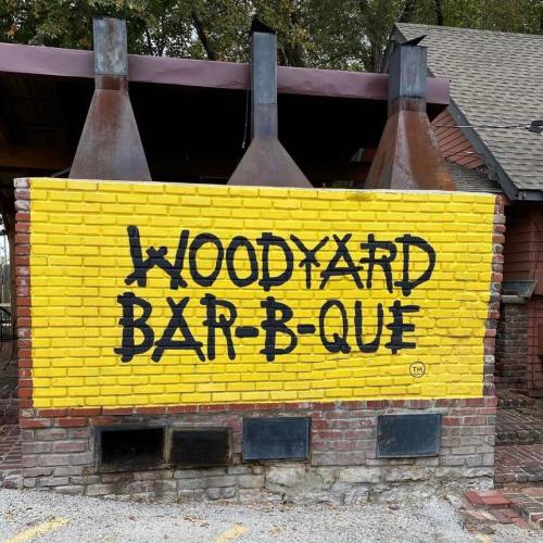 Bright yellow wall with the text "WOODYARD BAR-B-QUE" painted in bold black letters. Three metal chimneys above.