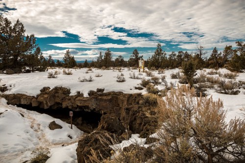 A snowy landscape with rocky terrain, sparse vegetation, and a cave entrance under a cloudy sky.