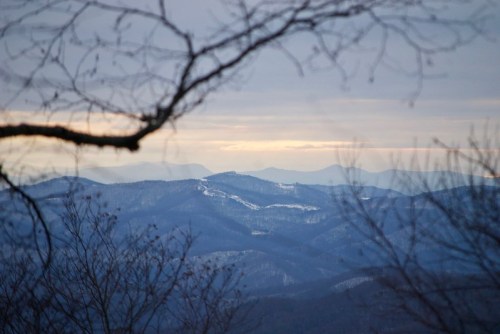 A scenic view of layered mountains under a cloudy sky, framed by bare tree branches in the foreground.