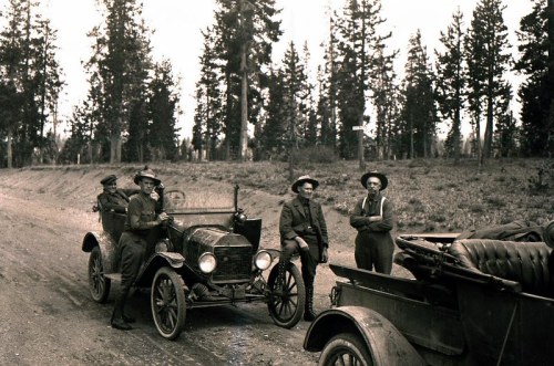 Four men in vintage clothing stand by early 20th-century cars on a dirt road, surrounded by tall trees.