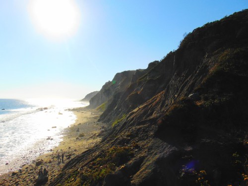 A sunlit coastal scene with cliffs, a sandy beach, and gentle waves under a clear blue sky.