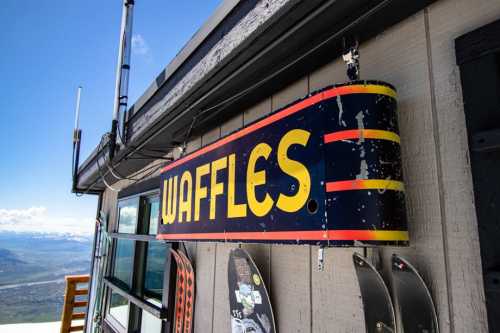 A colorful sign reading "WAFFLES" hangs outside a building with a scenic view in the background.