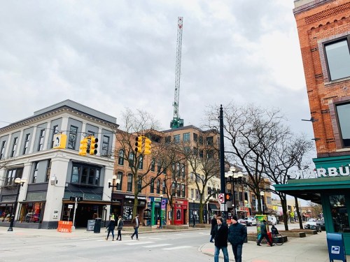 A city street scene with shops, traffic lights, and a tall crane in the background under a cloudy sky.