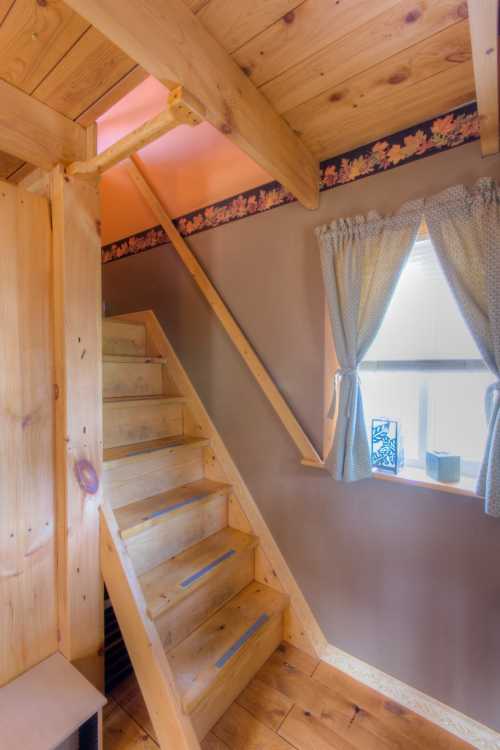 A wooden staircase leads to a cozy loft area, with a window dressed in light curtains and floral wallpaper.