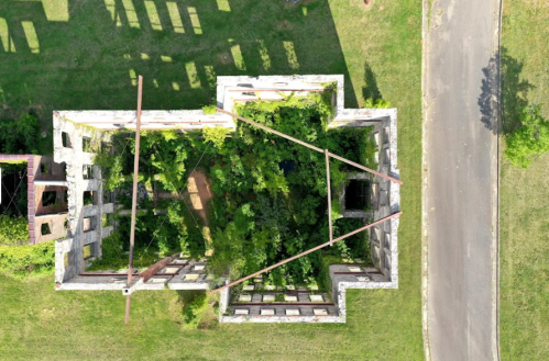 Aerial view of a dilapidated building overtaken by greenery, surrounded by grass and a nearby road.