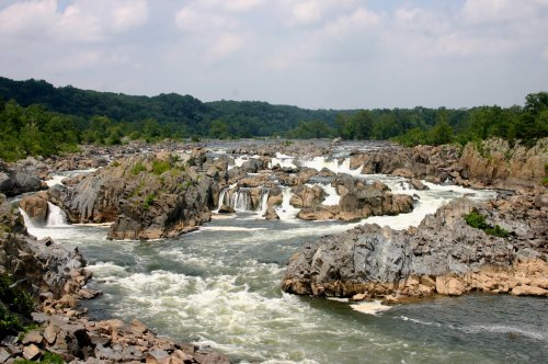 A scenic view of cascading waterfalls over rocky terrain, surrounded by lush greenery and a cloudy sky.