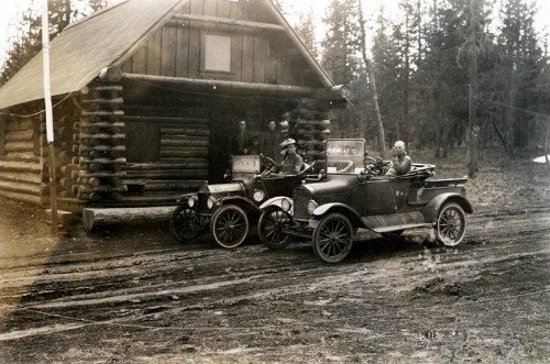 Two vintage cars parked in front of a log cabin, with people standing nearby in a forested area.