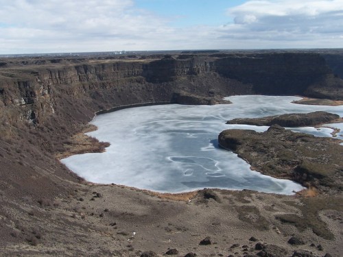 A frozen lake surrounded by rocky cliffs and sparse vegetation under a cloudy sky.