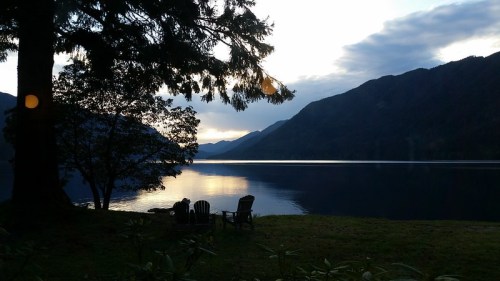 A serene lakeside scene at sunset, featuring chairs by the water and mountains in the background.