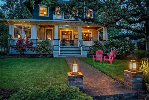 A charming blue house with a porch, surrounded by greenery, featuring two pink chairs and lanterns along a brick path.
