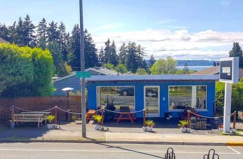 A small blue building with large windows, surrounded by greenery, overlooking a scenic view of water and trees.