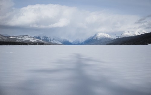 A serene winter landscape featuring a frozen lake surrounded by snow-covered mountains under a cloudy sky.