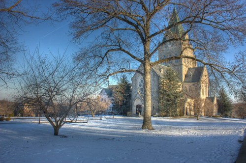 A snow-covered landscape featuring a large church with a tall steeple, surrounded by bare trees and a clear blue sky.