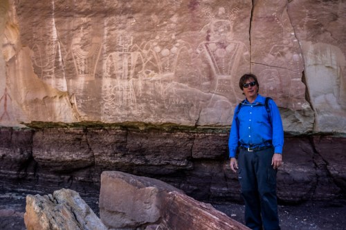 A person in a blue shirt stands near ancient rock art on a cliff face, surrounded by rocky terrain.