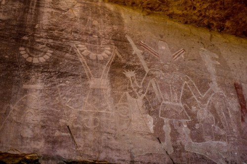 Ancient rock art depicting figures with elaborate attire and weapons on a textured stone surface.