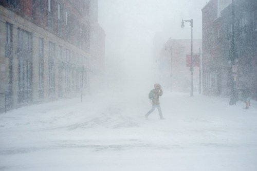 A person walks through a snowstorm in a deserted city street, visibility severely reduced by blowing snow.